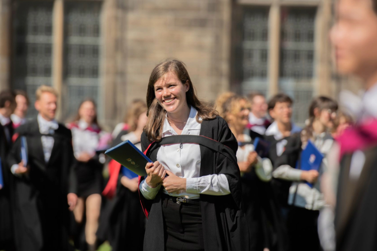 Female student smiling at graduation, holding graduation certificate, outside in sunshine